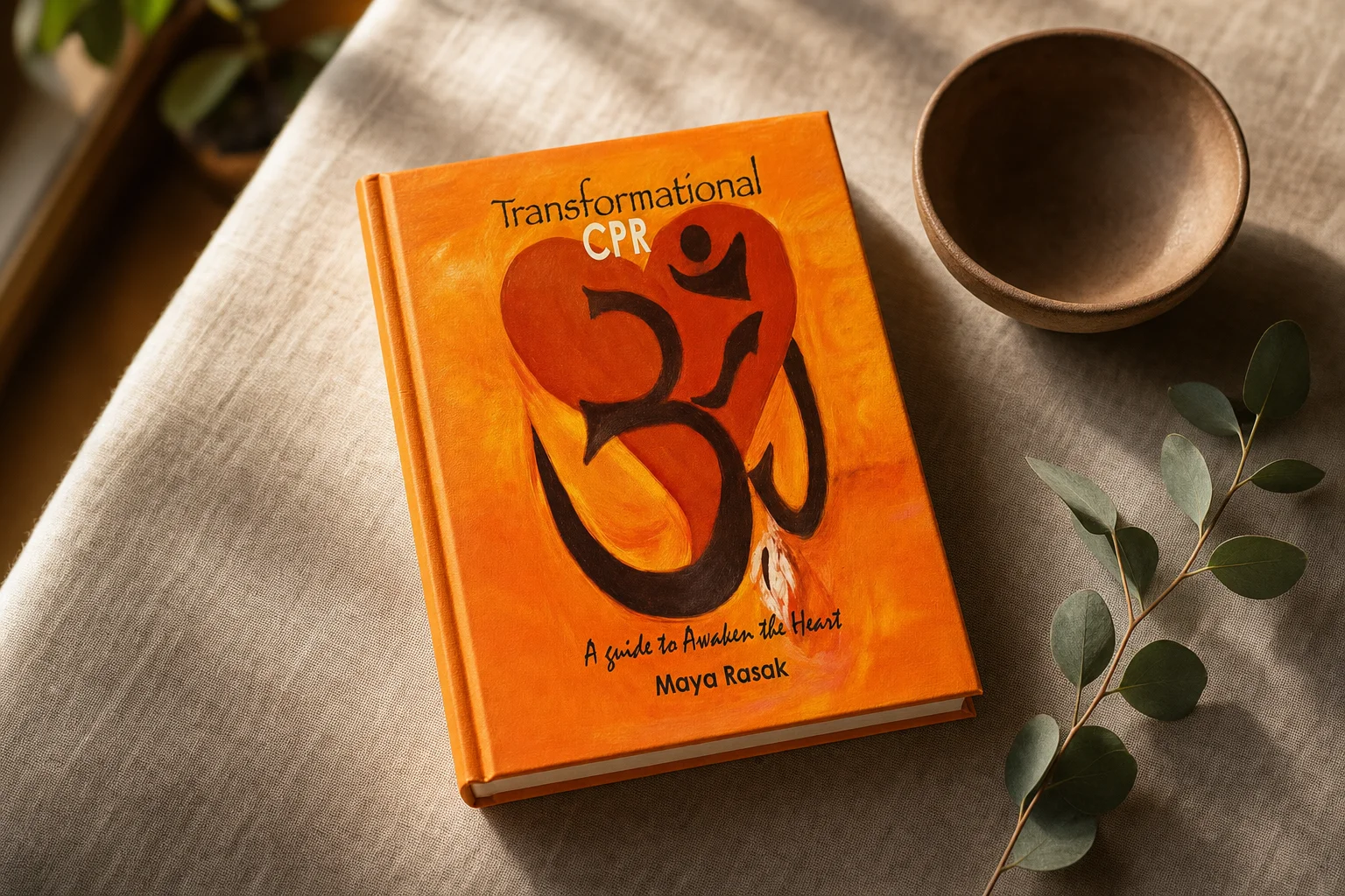 Transformational CPR: A Guide to Awaken the Heart by Maya Rasak, showing its orange painted cover with a heart and Om symbol, resting on linen beside a clay bowl and eucalyptus sprig